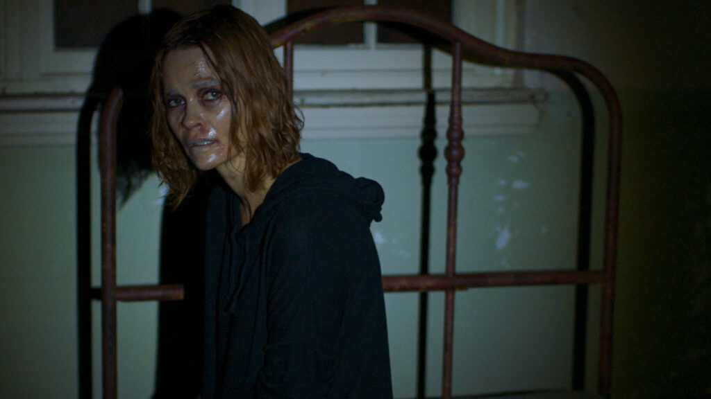 Nathalie Boltt als Angela sitz im Film Demonic auf einem Bett und schaut mit verhärmten Gesichtszügen in Richtung Kamera