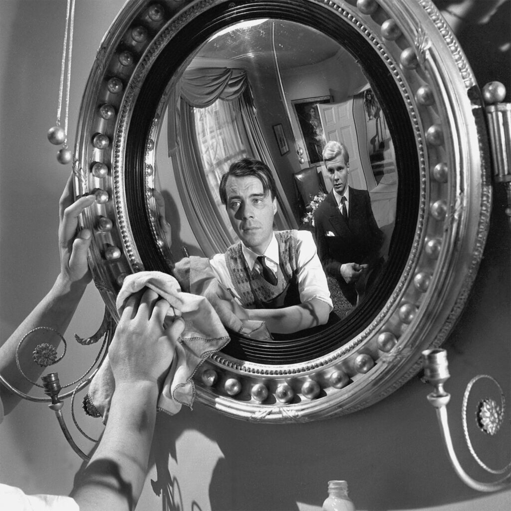 Während Hugo, gespielt von Dirk Bogarde, einen Wandspiegel putzt, spiegelt er sich mit Tony, gespielt von James Fox, darin.