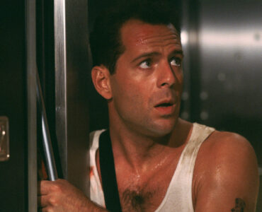 Bruce Willis ist im Film Stirb Langsam in einem Weißen Muskelshirt zu sehen und schaut dabei gespannt schräg nach oben