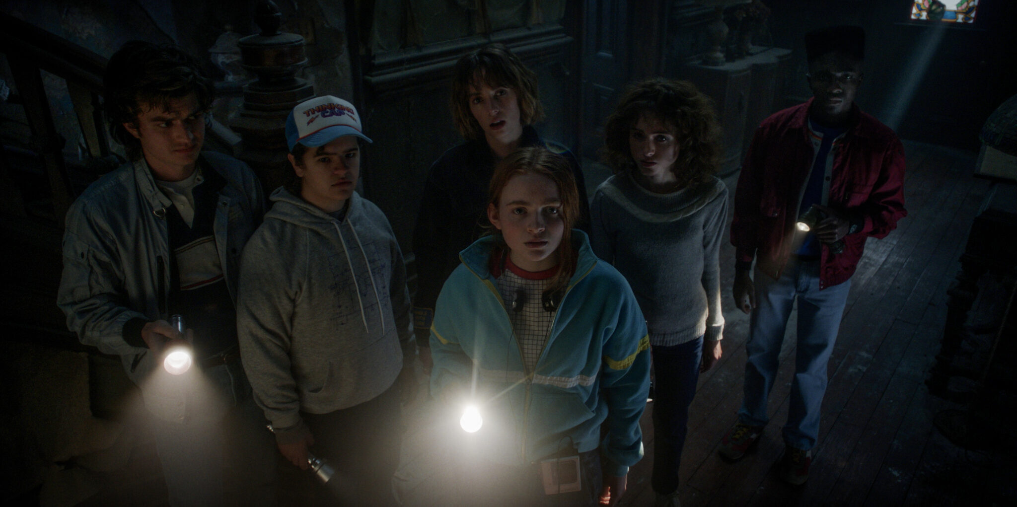 Man sieht in einem dunklen Raum sechs junge Menschen nebeneinander, drei davon haben Taschenlampen in der Hand.