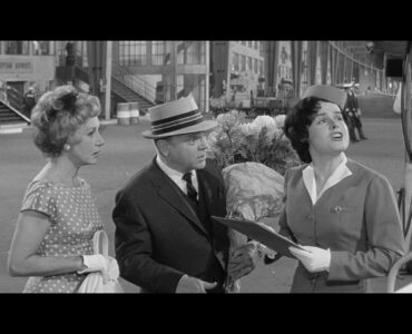 MacNamara (James Cagney) steht in Eins, zwei, drei mit seiner Frau und Blumenstrauß in der Hand am Flughafen.