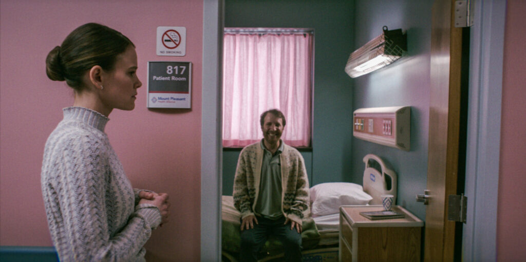 Ein Krankenhaus-Flur mit rosa Tapette. In einem Raum sitzt ein Mann in beiger Jacke, lächelt in die Kamera, aber ein diabolisches Lachen. Eine Szene aus dem Film Smile - Siehst du es auch?