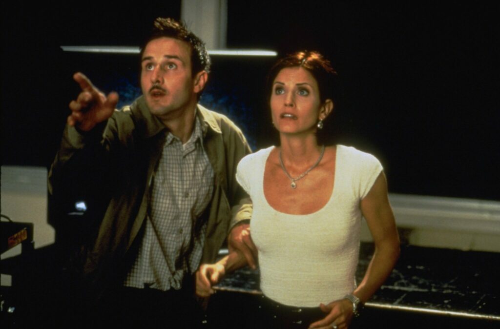 Eine Szene aus dem Film Scream 2: David Arquette als Dewey trägt eine beige-grüne Jacke und blickt mit erhobenen rechten Zeigefinger zu einem unbekannten Ziel. Rechts neben ihm steht Courtney Cox als Gale, sie trägt ein weißes, weit ausgeschnittenes Top und blickt erschrocken in die selbe Richtung.