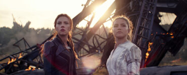 Natasha (Scarlett Johansson) und Yelena (Florence Pugh) stehen vor einem brennenden Wrack. Natasha hat rote Haare und trägt ein dunkles Outfit, Yelena ein weißes Kostüm.