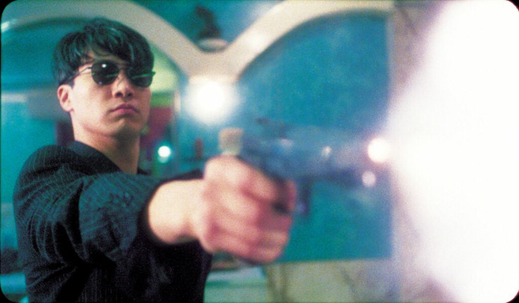Fallen Angels Hauptcharakter Wong Chi-Ming steht am linken Rand und feuert mit ausgestrecktem Arm eine Hamdfeuerwaffe nach rechts vorne. Er trägt eine Sonnenbrille. Von hinten wird er von einer Lampe angeleuchtet, die sich in einem Spiegel spiegelt. Rechts im Bild ist der grelle Blitz des Mündungsfeuers.