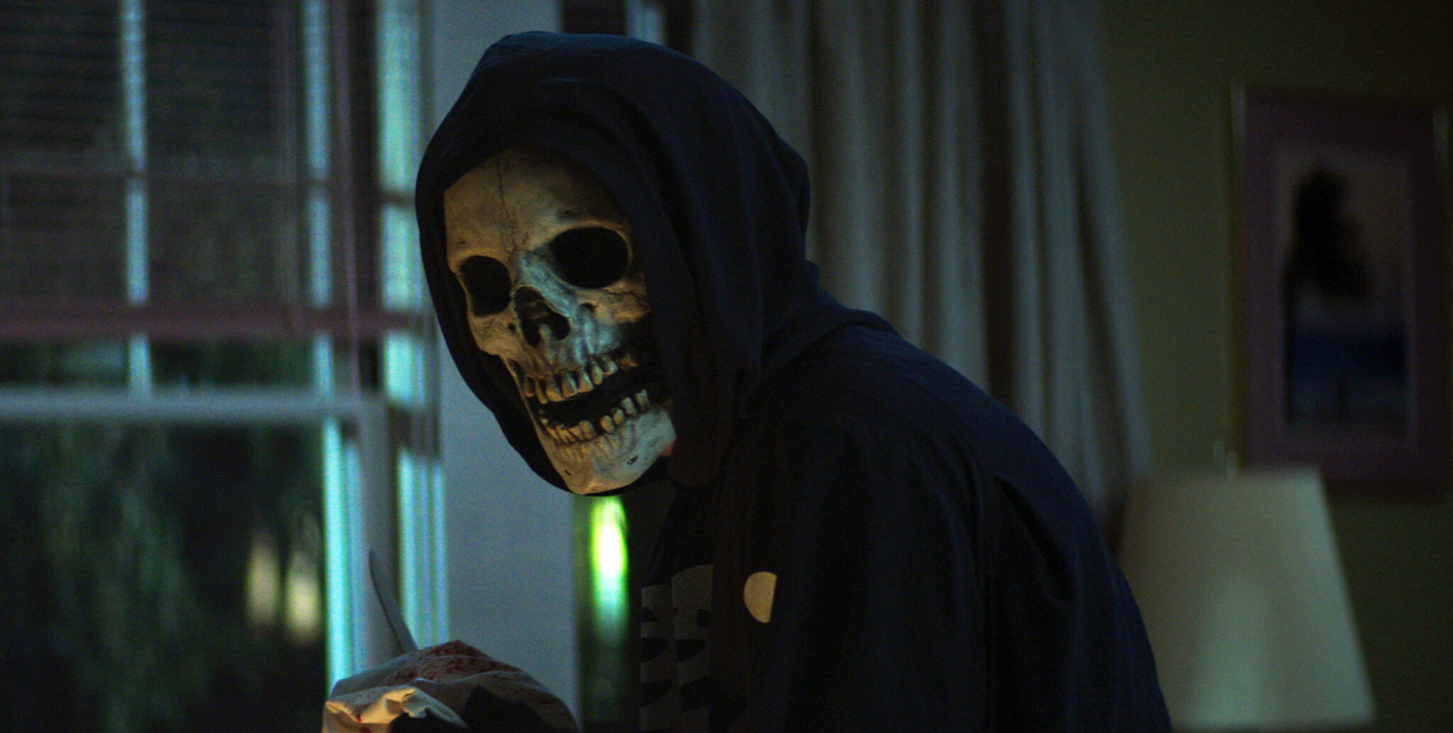 Eine Person mit Totenkopfmaske und Kapuzenpulli in schwarz in einem dunklen Zimmer. Die Person hält ein Messer in der Hand.