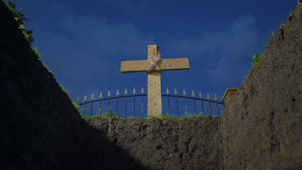 Aus einem ausgehobenen Grab ist das hölzerne Kreuz eben jenes zu sehen. Dahinter ein Zaun und der Himmel. Nobody Loves You and You Don't Deserve to Exist 
