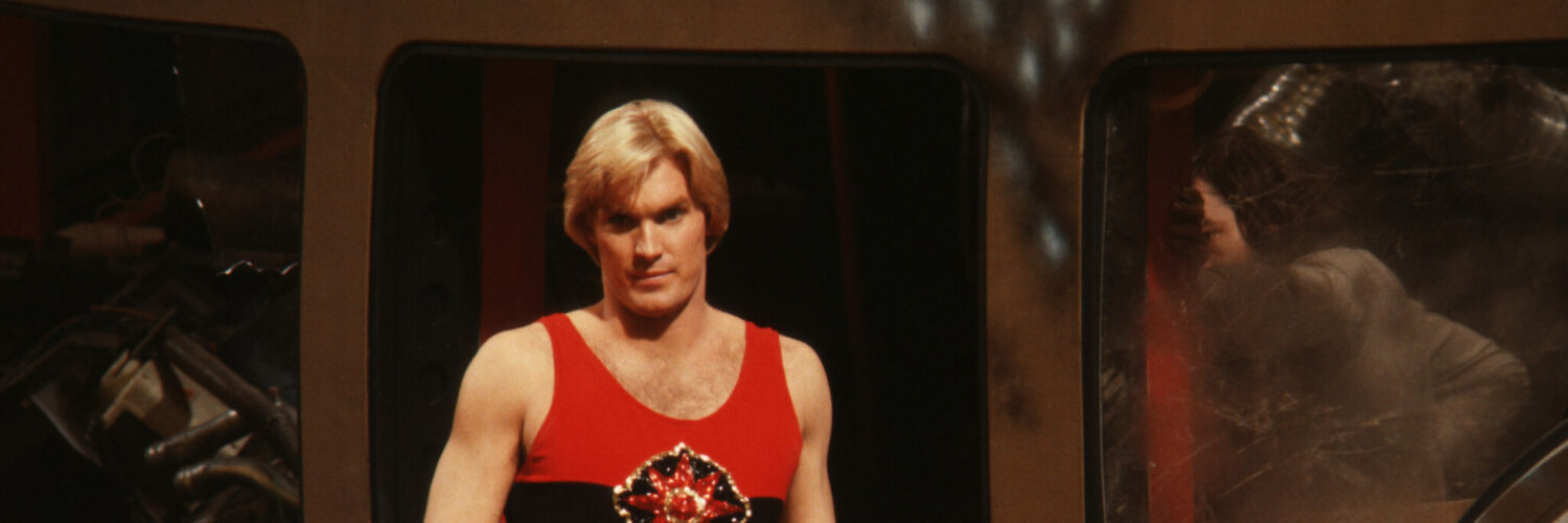 Flash Gordon stehe in ärmellosen schwarz-roten Shirt vor einigen Trümmern. In seiner rechten Hand hält er ein goldenes Schwert und blickt auf etwas nach unten.