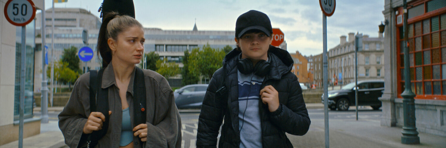 Flora und Max gehen nebeneinander auf der Straße in Dublin. Sie trägt eine Gitarre auf dem Rücken und schaut zu ihrem Sohn rüber