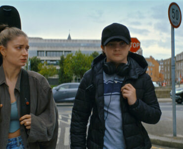 Flora und Max gehen nebeneinander auf der Straße in Dublin. Sie trägt eine Gitarre auf dem Rücken und schaut zu ihrem Sohn rüber