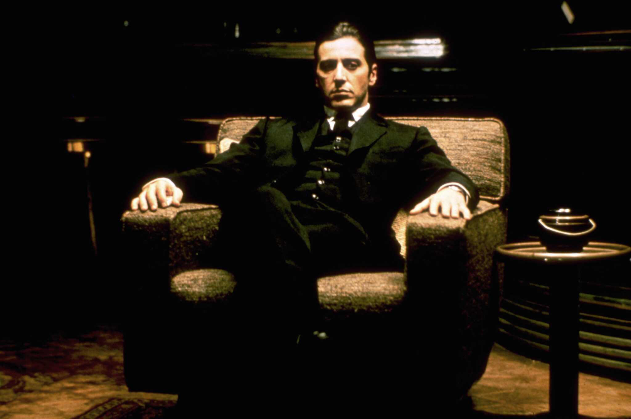 Auf dem Bild erkennt man Michael Corleone im Anzug, wie er selbstbewusst auf einem Sessel sitzt - Der Pate-Trilogie