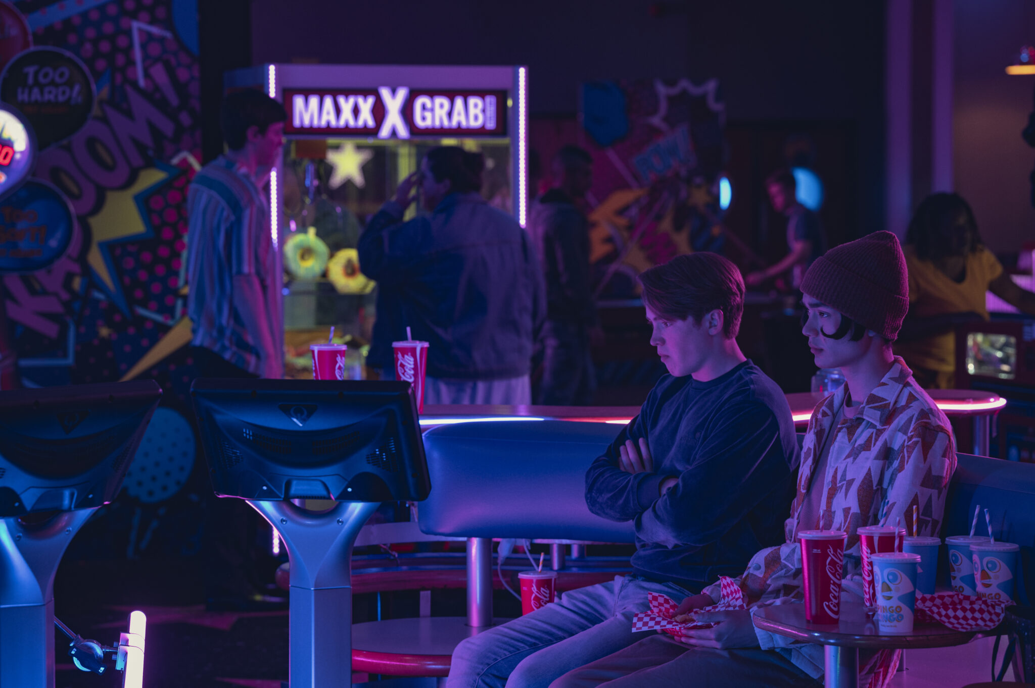 Eine neonbeleuchtete Spielhalle. Vorne recht sitzen zwei Jungen mit nachdenklichen Gesichtern und zögerlicher Körperhaltung.