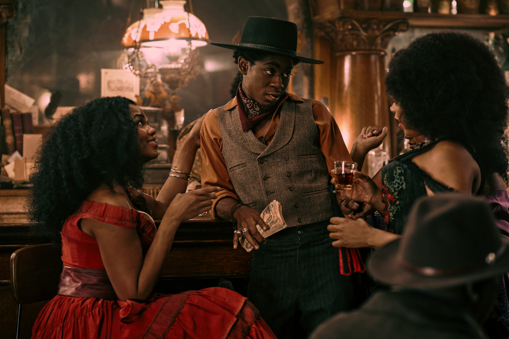 RJ Cyler als Jim Beckwourth zwischen zwei nobel gekleideten Damen an der Theke im Saloon. Er hält ein Bündel Geldscheine in der Hand und trägt einen schwarzen Hut.