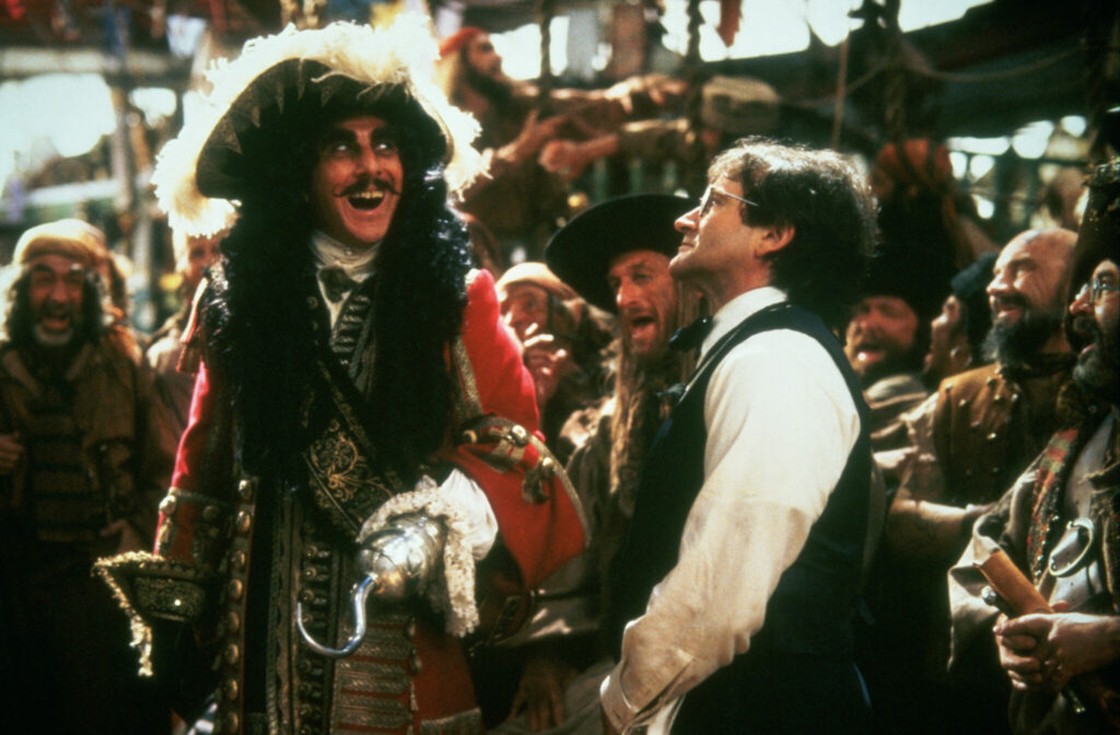 Zu sehen ist eine Szene aus "Hook", in der Dustin Hoffman als Captain Hook auf dem Deck seines Schiffs steht und den gefesselten Peter (Robin Williams) begrüßt. Dabei blickt der Bösewicht manisch freudig über seinen Gefangenen hinweg zu den Seinen, während Peter nur mit ernster Miene auf Hook blickt. - Robin Williams Filme