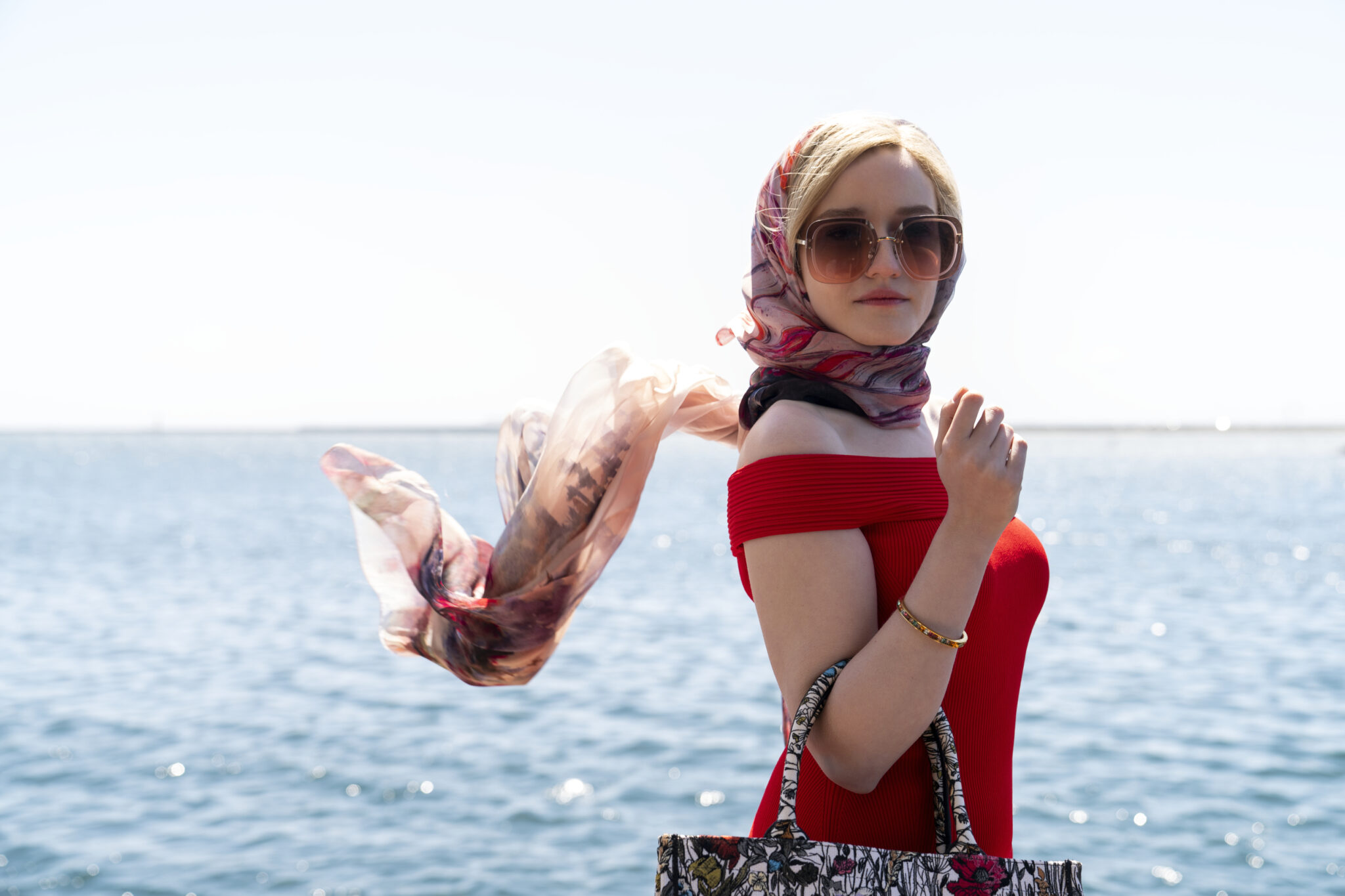 Julia Garner mit rotem Kleid vor einem Meer im Hintergrund, sie trägt außerdem ein Kopftuch und eine große Sonnenbrille.