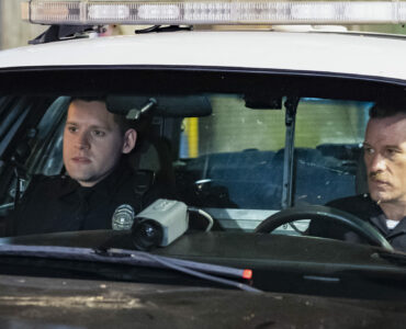 Nick Holland und Ray Mandel sitzen in ihrem Streifenwagen. Auf der Armatur liegt eine große Kamera die nach vorne filmt. Beide Polizisten ziehe eine ernste Miene.