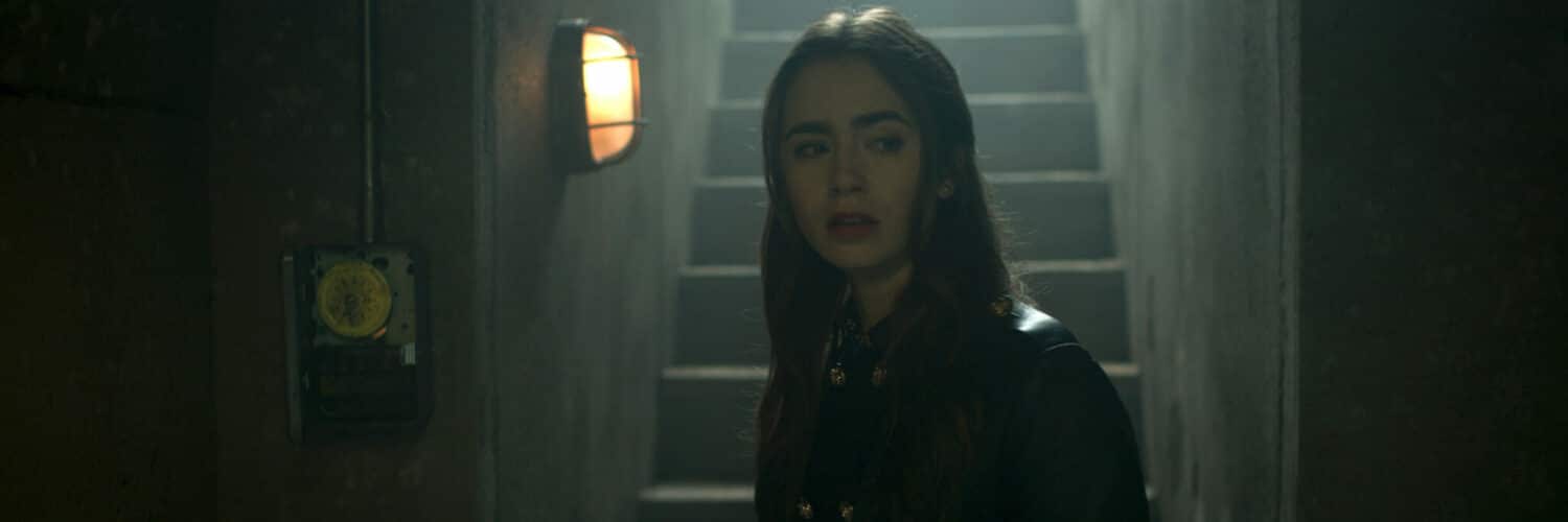 Lily Collins spielt die Hauptfigur Lauren Monroe in "Inheritance". Auf dem Bild geht diese zögerlich durch einen dunklen Korridor. Im Hintergrund ist eine Treppe zu sehen über die sie vermutlich runter in diesen Korridor gegangen ist.