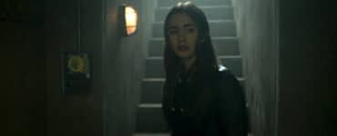 Lily Collins spielt die Hauptfigur Lauren Monroe in "Inheritance". Auf dem Bild geht diese zögerlich durch einen dunklen Korridor. Im Hintergrund ist eine Treppe zu sehen über die sie vermutlich runter in diesen Korridor gegangen ist.