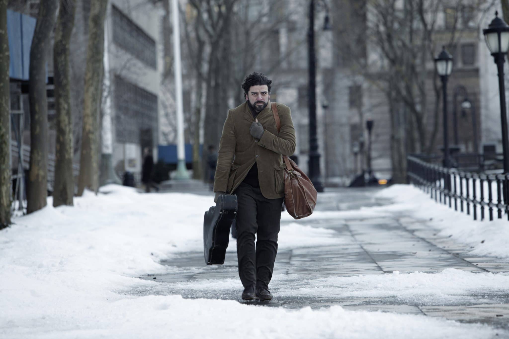 Llewyn Davis (Oscar Isaac) läuft mit einem Gitarrenkoffer in der Hand allein auf einem verschneiten Gehweg in einer Großstadt.