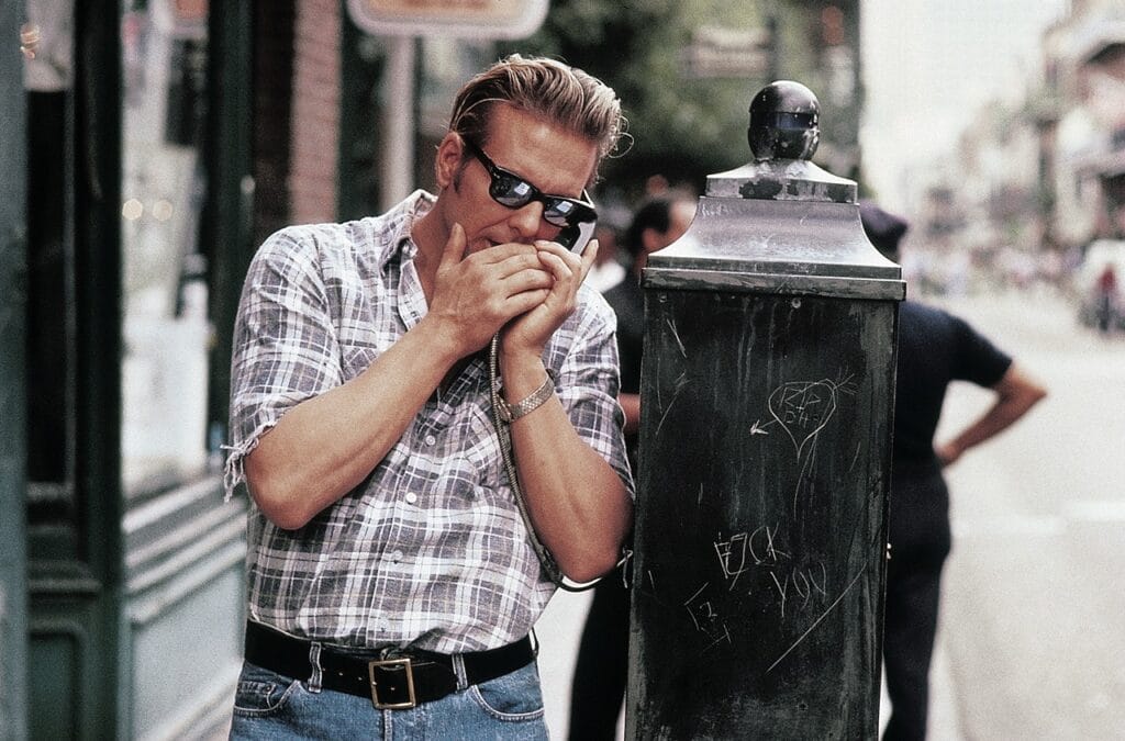 Johnny (Mickey Rourke) steht an einem öffentlichen Telefon und hält sich den Hörer dicht ans Ohr. Er trägt Sonnenbrille und hält sich die freie Hand vor den Mund.