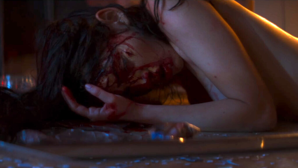 Eli liegt mit blutverschmiertem Gesicht auf den Scherben eines zerbrochenen Glasstisches - Jezabel