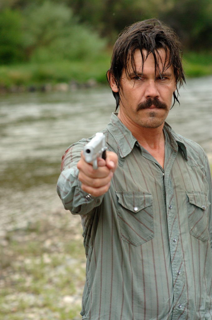 Josh Brolin als Llewlyn Moss, durchnässt an einem Fluss, richtet eine Waffe auf sein Gegenüber