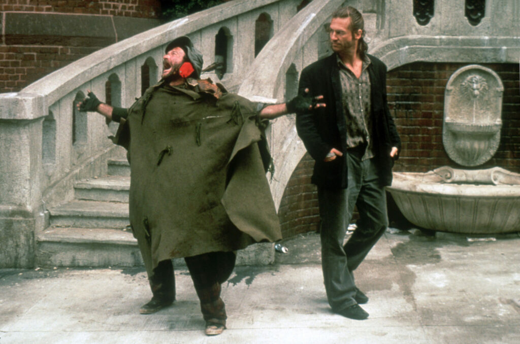 Zu sehen ist eine Szene aus "König der Fischer", in der Robin Williams und Jeff Bridges auf der Straße vor einer Treppe stehen. Williams trägt verlumpte Kleidung und streckt die Arme von sich weg, während er den Mund weit geöffnet hat. Es wirkt als wenn er freudig singt. Jeff Bridges steht zu seiner Linken und schaut Williams an, während er die Hände in der Tasche hat. - Robin Williams Filme 