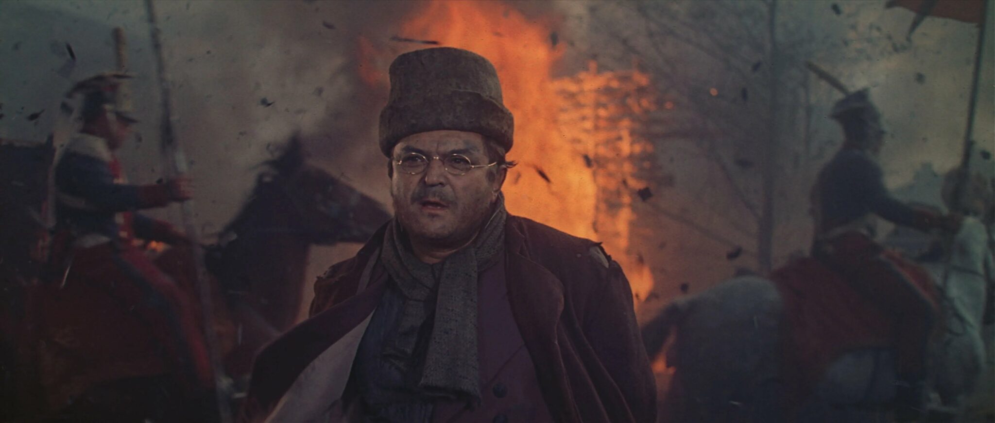 Pierre Bezukhov (Sergei Bondartschuk) steht in Mitten von Trümmern im verschneiten sowie verwüsteten Moskau. Berittene Soldaten reiten hinter ihm und es brennt.