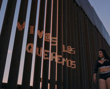 Lady Candy läuft mit ernster Miene an ein Gitter entlang mit der Aufschrift "Viva Las Queremos"