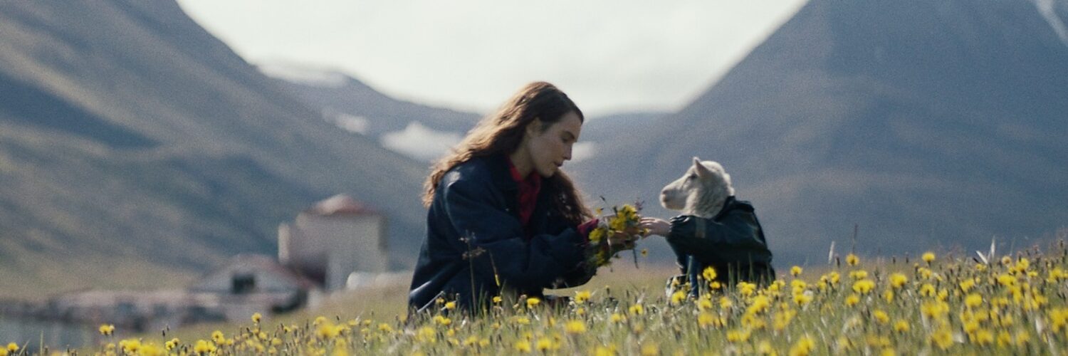 Eine Wiese im Vordergrund, im Hintergrund Berge. Auf der Wiese sitzt die Schauspielerin Noomi Rapace mit einem Schaf. Ein Szenenbild aus dem Film Lamb