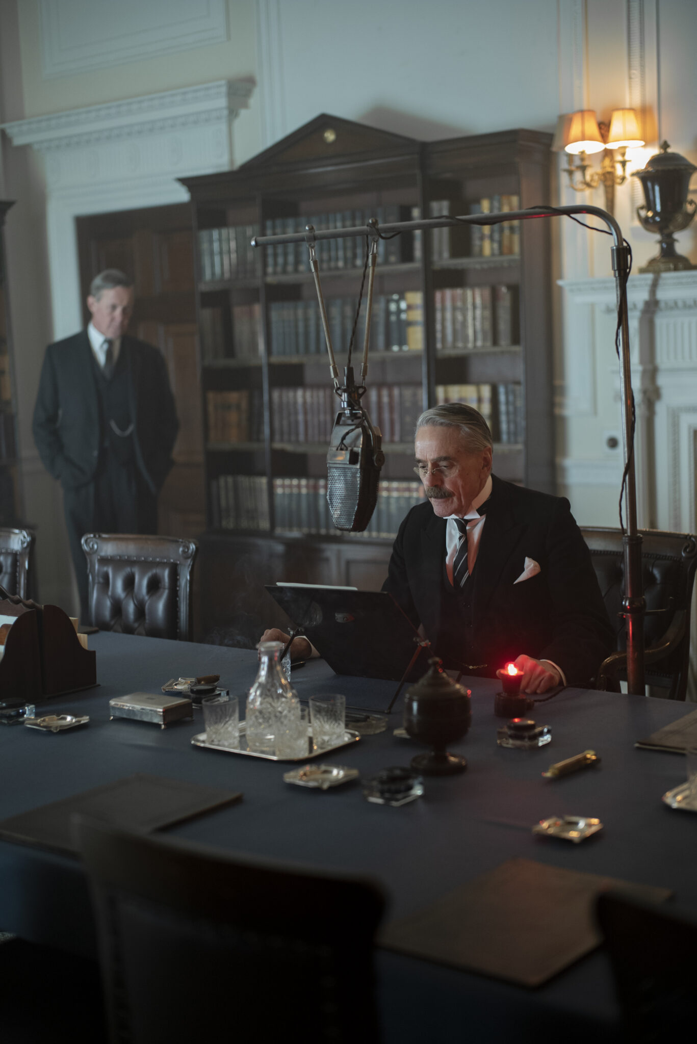 Jeremy Irons als Chamberlain liest vor einem Mikrofon sitzend von einem Blatt ab. Im Hintergrund sieht man einen Mann im Anzug und eine Bücherwand. München - Im Angesicht des Krieges