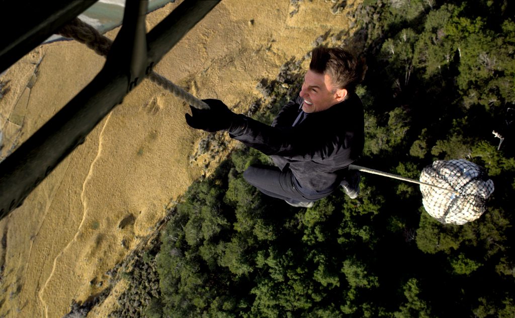 Tom Cruise mit beiden Händen an einem Seil, das an einem Helikopter befestigt ist, der gerade abhebt.