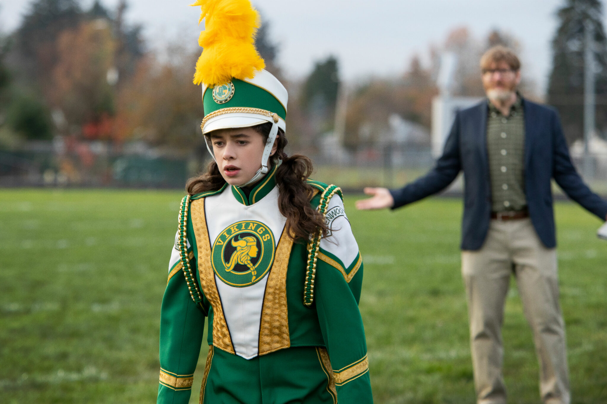 Emily (Isis Hainsworth) in einem grünweißgoldenen Kapell-Outfit samt aufwändigem Helm auf einer offenen Wiese. Hinten unscharf ein erwachsener Mann mit beiger Hose und blauem Blazer.