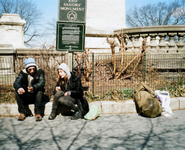 Mike (Buddy Duress) und Harley (Arielle Holmes) sitzen rauchend am Straßenrand. Rechts neben ihnen liegt der Rucksack von Harley und einige Plastiktüten. Im Hintergrund befindet sich das Soldiers' and Sailors' Monument in New York. © Koch Films