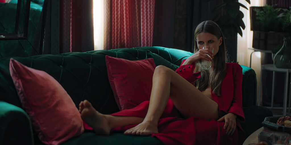 Mara, gespielt von Aleksandra Revenko, liegt in lasziver Pose, von ihrem roten Kleid nur unzureichend verhüllt, auf ihrer Couch.