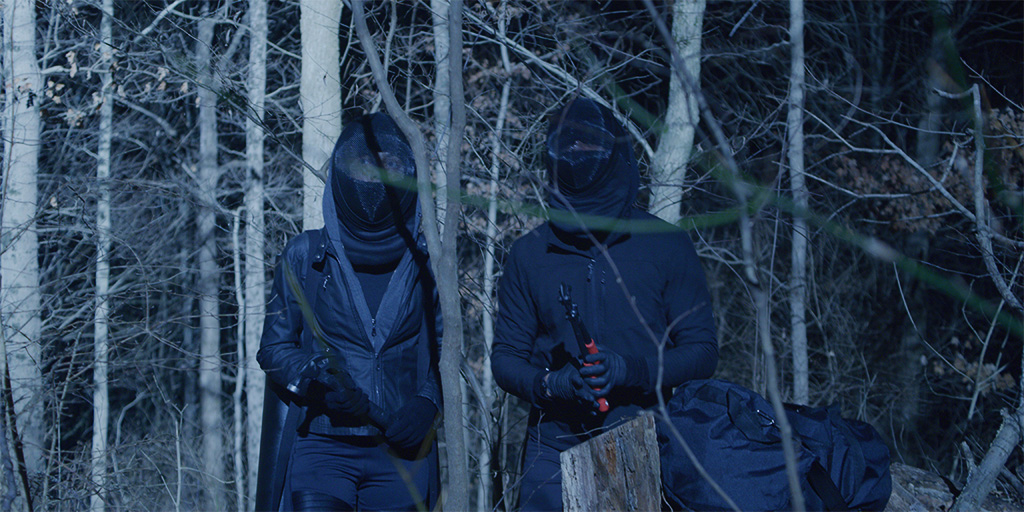 Zwei komplett vermummte Personen stehen mit Werkzeug in der Hand im dunklen Wald. 