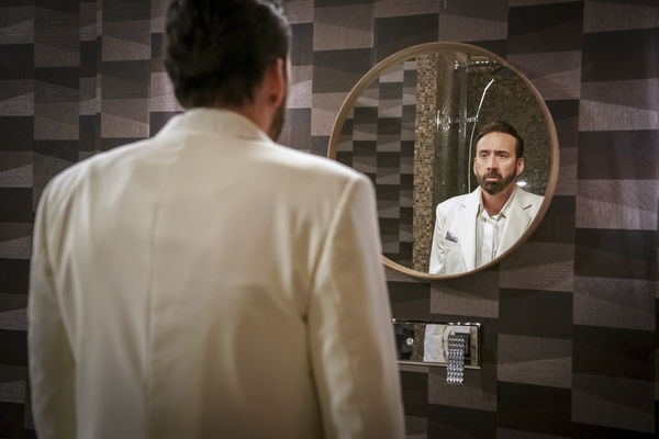In einem Badezimmer voller grauer und weißer Kacheln, steht Nicolas Cage vor einem Spiegel und betrachtet sein Spiegelbild.
