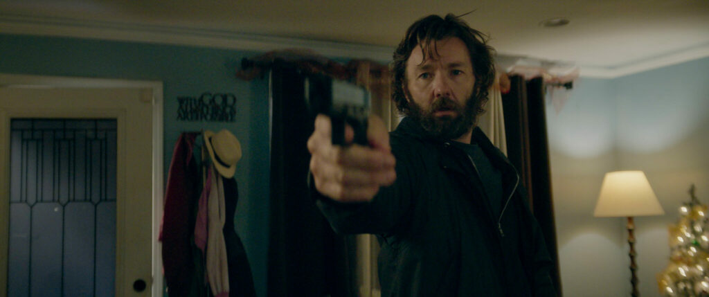 Ein Mann mit Vollbart und lockigen Haaren richtet eine Waffe auf ein unbekanntes Ziel. Eine Szene aus dem Film Master Gardener.