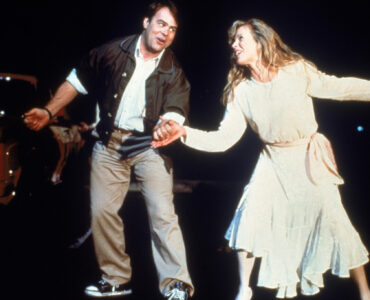 Dan Aykroyd und Kim Basinger halten sich die Hände und scheinen zu tanzen und zu singen. - "Meine Stiefmutter ist ein Alien"