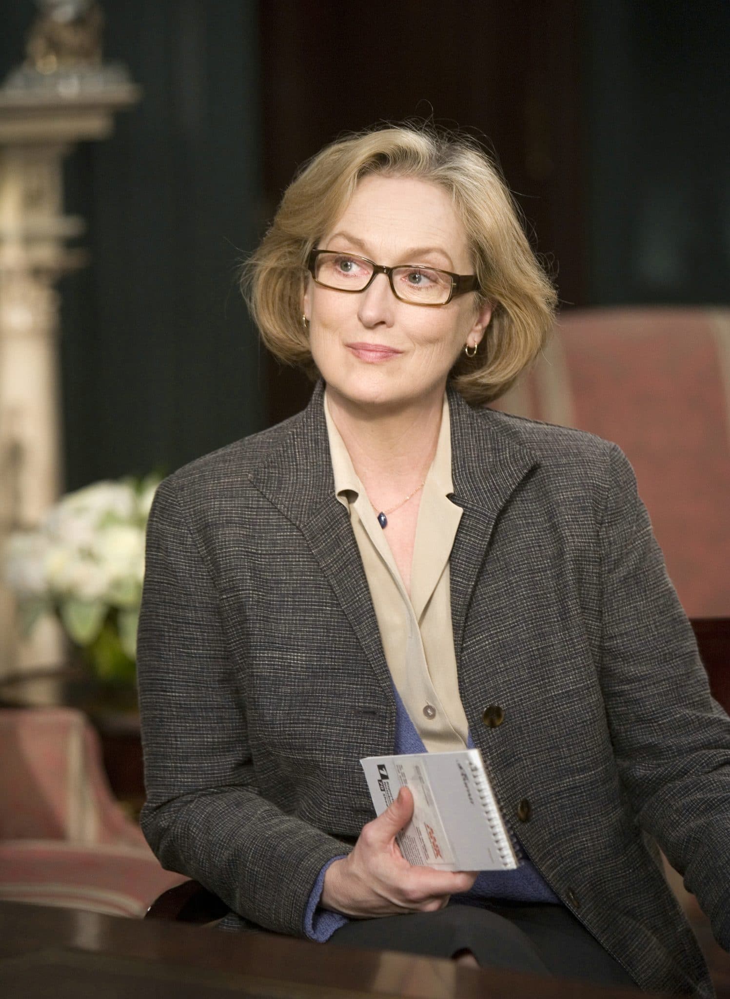 Meryl Streep in einem Businessoutfit mit Brille und einem Schreibheft in der Hand.