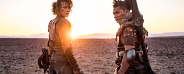Milla Jovovich & Tony Jaa stehen in ihren Jägermonturen Seite an Seite in einer kargen Ödnis. Beide blicken über ihre Schultern hinter sich, während im Hintergrund die Sonne hinterm Horizont verschwindet. - "Monster Hunter"