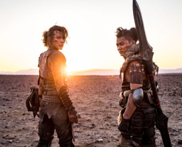 Milla Jovovich & Tony Jaa stehen in ihren Jägermonturen Seite an Seite in einer kargen Ödnis. Beide blicken über ihre Schultern hinter sich, während im Hintergrund die Sonne hinterm Horizont verschwindet. - "Monster Hunter"