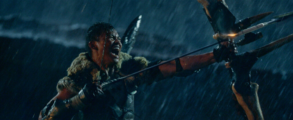 Martial Arts-Star Tony Jaa ist hier in seiner Jägermontur mit leichter Panzerung und Fell über den Schultern zu sehen. In scheinbar strömendem Regen spannt er gerade einen Pfeil mit seinem übergroßen Bogen und blickt dabei brüllend auf sein Ziel. - "Monster Hunter"
