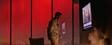Richard steht in Unterwäsche vor seinem Kamin, über dem ein großes TV-Gerät hängt, im Hintergrund sieht man den nächtlichen Blick auf Manhattan vor dem Abendrot - Mosquito State