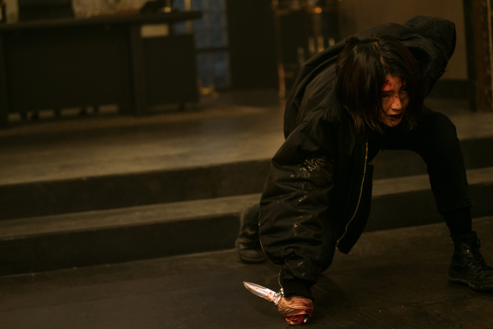 Ein junges Mädchen in gebückter, kampfbereiter Haltung in schwarzem Outfit vor einer Treppe. Sie hat ein blutiges Messer in der Hand.