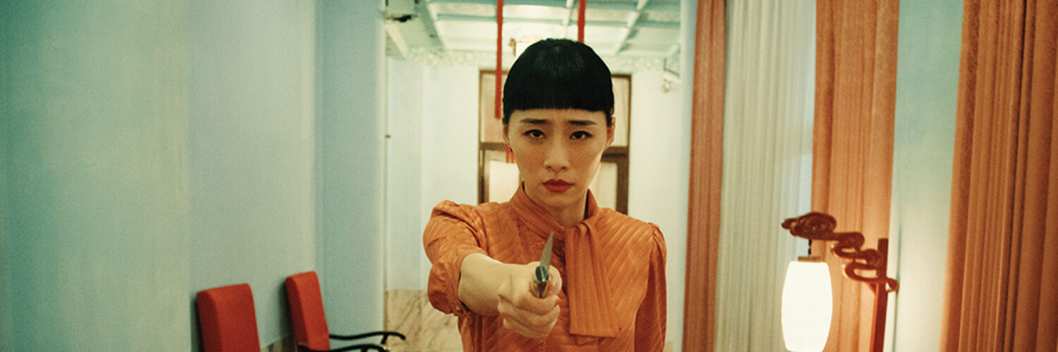 Nina Wu (Wu Ke-Xi) läuft mit starren Blick direkt in die Kamera gerichtet in orangenfarbenen Kleid und gezücktem Messer einen engen Gang entlang.
