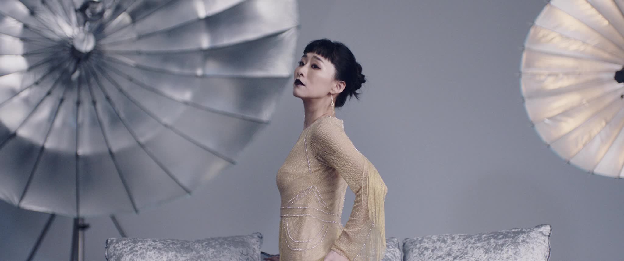 Nina Wu (Wu Ke-Xi) posiert mit angestrengter Miene, dunklem Lippenstift und in beigem Kleid in einem grau hinterlegten Fotostudio.