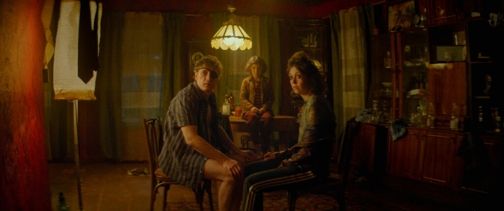 Viktoriya Korotkova als Olga, Sofya Krugova als Masha und Aleksandr Yatsenko als Oleg sitzen im Film No Looking Back an einem Tisch in einem düsteren Wohnzimmer und blicken fragend in die Kamera.