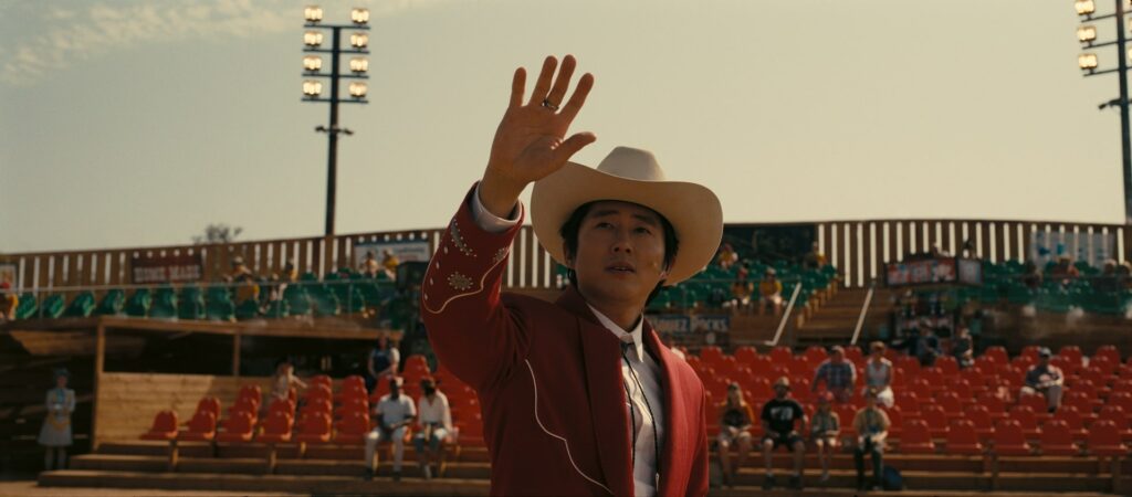Ricky 'Jupe' Park (Steven Yeun) schaut über die Kamera, trägt einen roten Anzug und einen weißen Western-Hut. Im Hintergrund sind die Tribünen seines Western-Themenparks zu sehen in Nope.