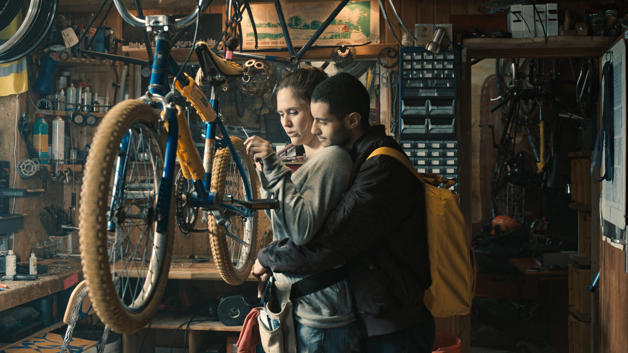 Karim umarmt Lilly in Nur ein Augenblick von hinten, während sie in ihrer Werkstatt gerade ein Fahrrad repariert.